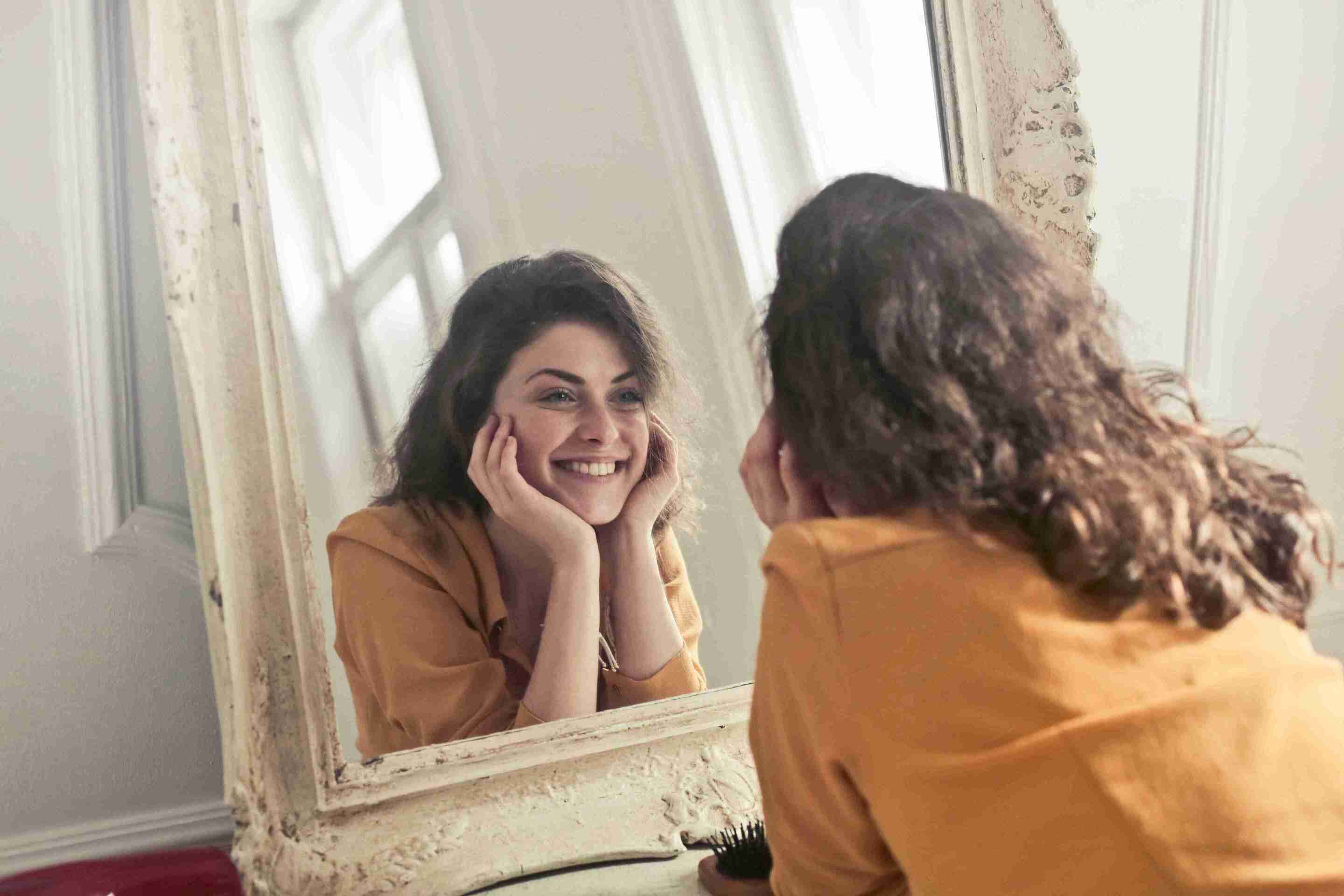Woman smiling at her reflection in the mirror, symbolizing confidence and self-care due to the use of rawnaqy.com products.